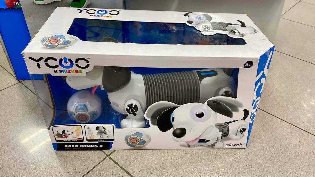 Roboterhund im Test: ein Yoco Robo Dackel in weiß-grauem Design befindet sich in seinem Karton mit Sichtfenster auf einem beigefarbenen Fliesenboden.