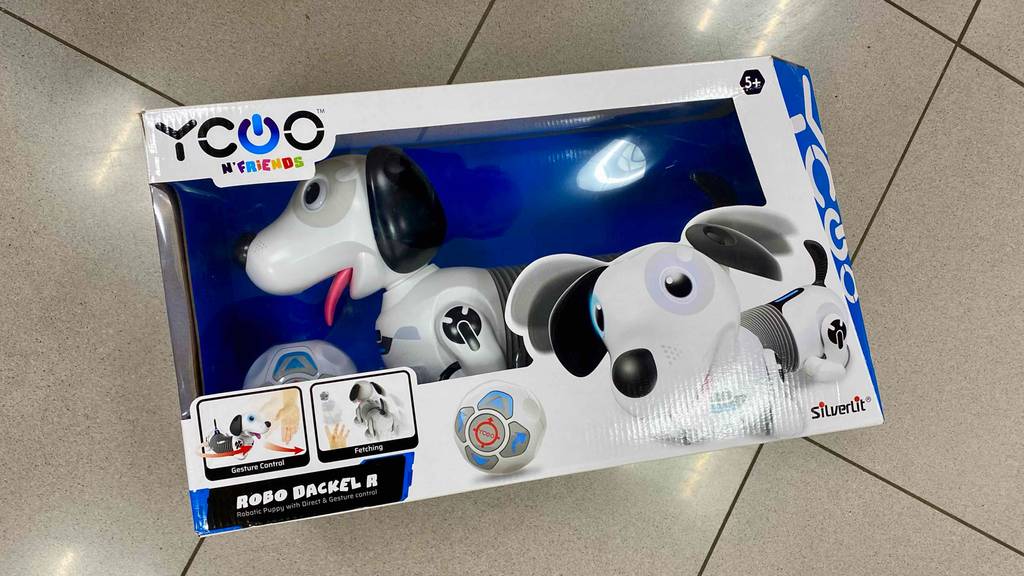 Roboterhunde-Test: ein Yoco Robo Dackel im weiß-grauen Design liegt in seinem Karton inklusive Sichtfenster auf beigefarbenen Fliesen.