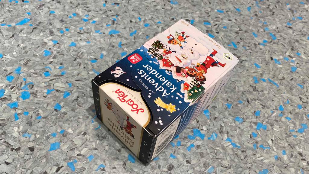 Ein getesteter Adventskalender der Marke Yogi Tea liegt mit Blick auf die Entnahme-Vorrichtung auf einer blau-grau gesprenkeltem Oberfläche.