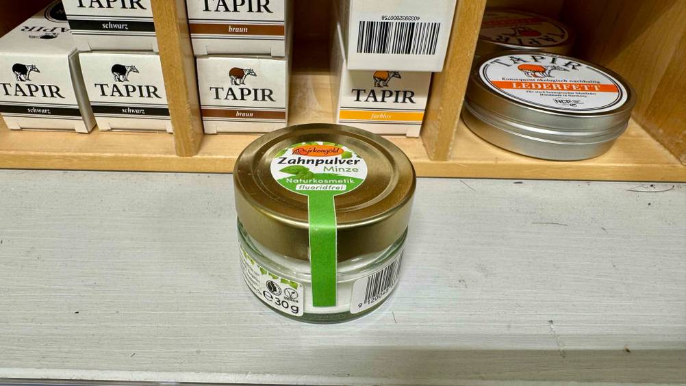 Eine Dose des getesteten Zahnpulvers von schräg oben vor einem Regal mit Produkten.