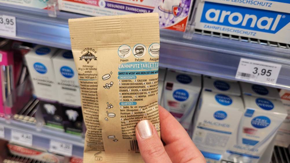 Eine Hand hält eine Packung der getesteten Zahnputztabletten mit der Rückseite nach vorne vor ein Regal.