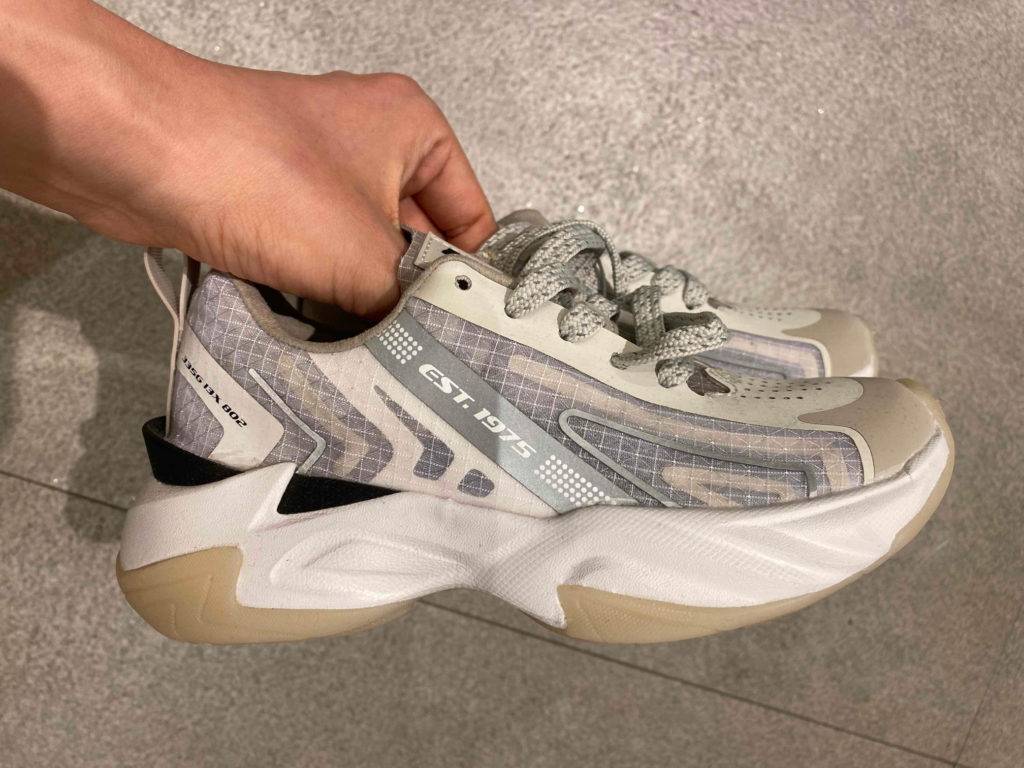 Seitenansicht weißer Laufschuhe für Kinder im Test mit grauen Design-Elementen in einer Hand vor grauen Fliesen.