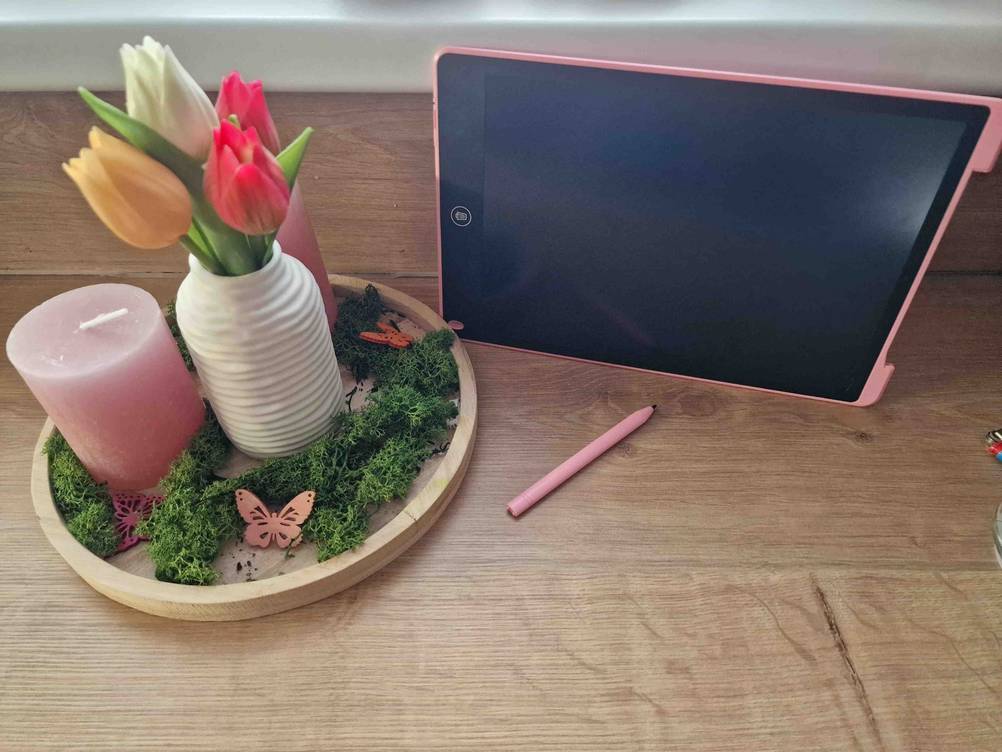 Rosa Zaubertafel im Test inklusive optischem Stift befinden sich auf einem Tisch neben einem Blumen-Kerzen-Gesteck.