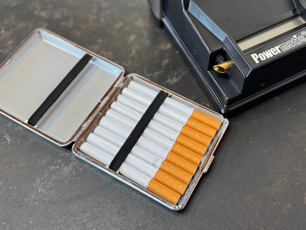 Ein Etui mit Zigaretten, im Hintergrund die getestete Zigarettenstopfmaschine von Powermatic.