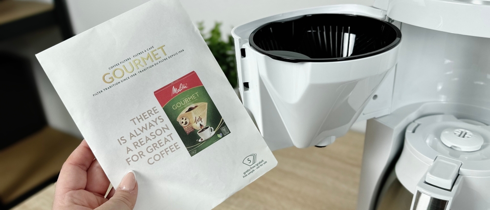 Eine Hand hält ein Paket Melitta-Gourmet-Filtertüten. Die Maschine mit geöffnetem Schwenkfilter ist im Hintergrund zu sehen.