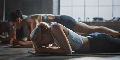 3 Personen in Sportkleidung in einem Trainingsraum. Die Frau ganz vorn liegt erschöpft auf dem Bauch, den Kopf hat sie auf den Händen abgestützt. Die Personen im Hintergrund befinden sich in Liegestützposition.