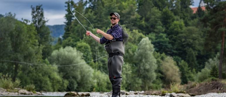 Angler mit Wathose und Fliegenrute im Wasser beim Angeln bei Sonne im klaren Fluss stehend und werfend